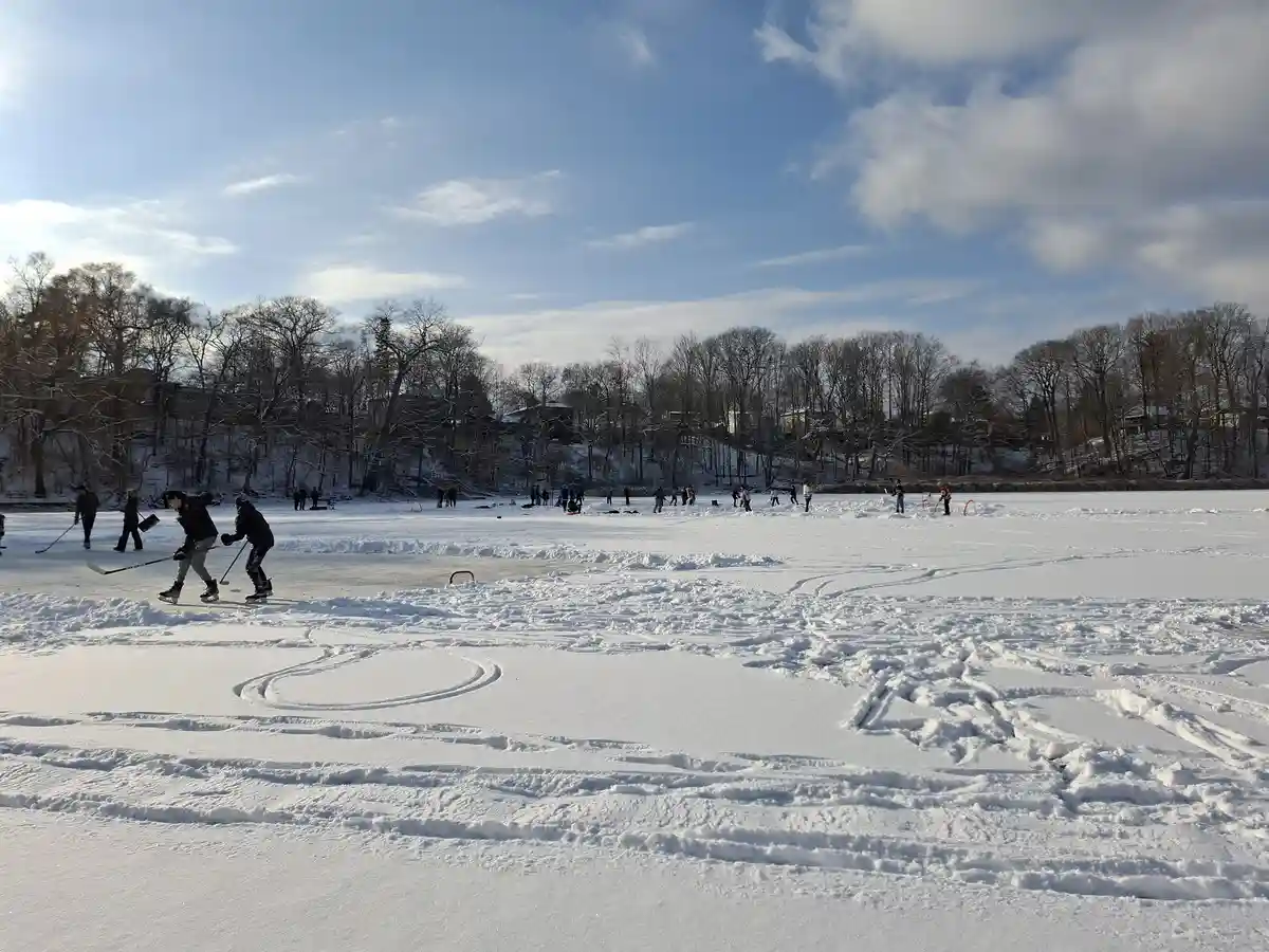 A wide shot of a frozen lake where people are playing ice hockey on the ice, with snow-covered ground in the foreground…