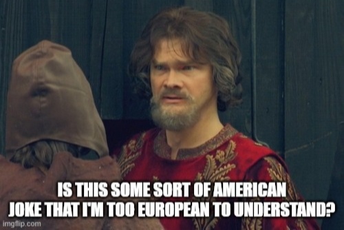 A man with a beard and curly hair wearing a red embroidered tunic looks confusedly at another person wearing a brown hooded…