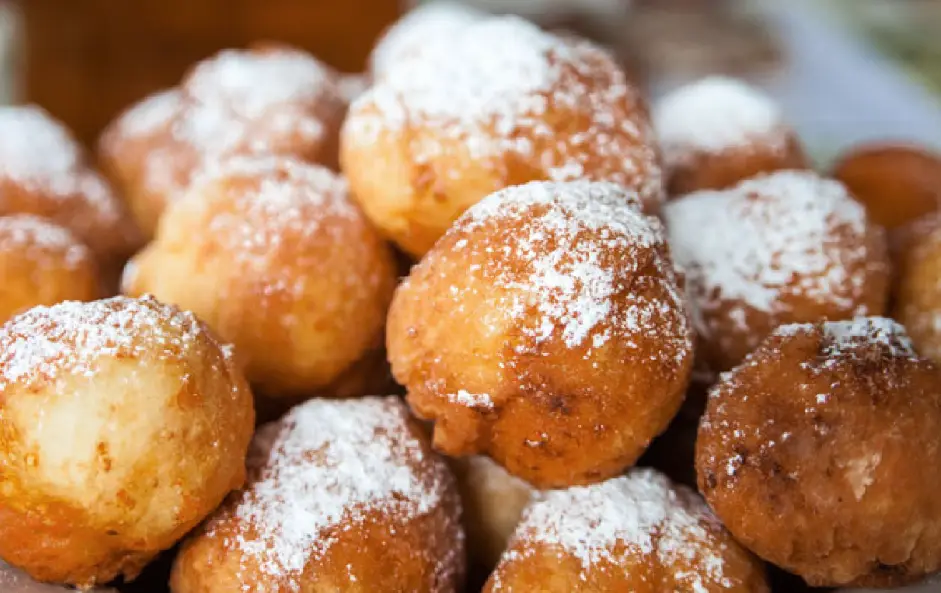 A close-up photo shows a pile of golden-brown, spherical fried pastries dusted with white powdered sugar. Overlaid text…