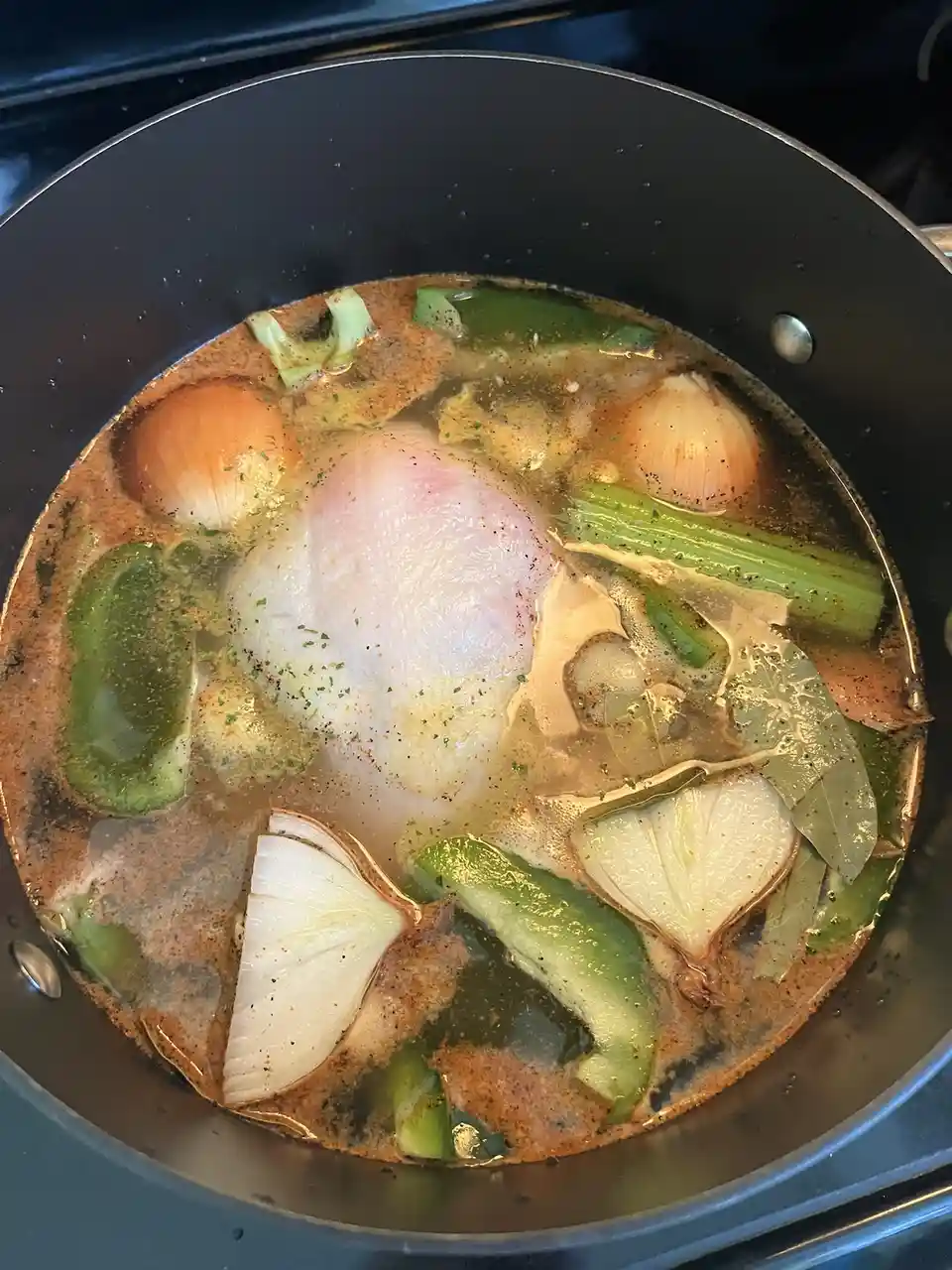 A top-down view of a black pot on a stovetop containing a simmering broth with a whole raw chicken breast, sliced onions…