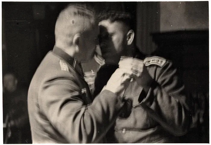 A black-and-white historical photograph shows two men in military uniforms, one older with a shaved head and the other…