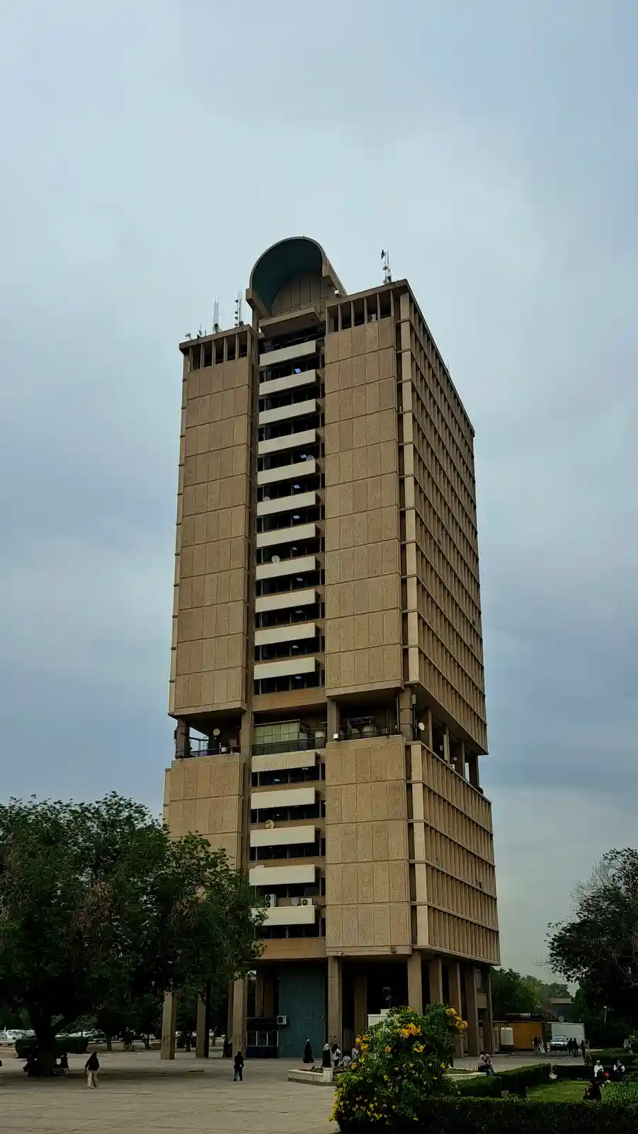 a tall, beige, brutalist-style building with a distinctive curved top section stands against a cloudy sky, surrounded by…