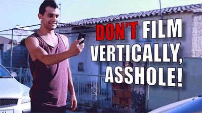 A man in a purple tank top holds a phone while standing outdoors near cars and a fence. Another person repeatedly reaches…