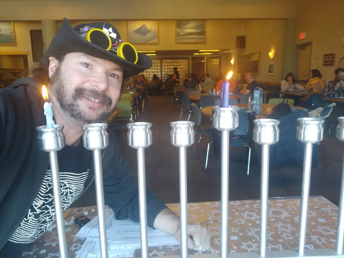 a man with a beard and a black hat adorned with yellow goggles smiles at the camera while sitting at a table with a menorah…