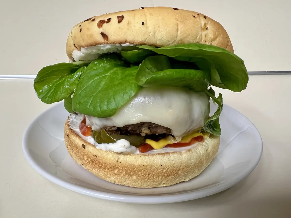 A close-up photo of a burger on a white plate, featuring a toasted bun with visible seeds, a beef patty, melted white…