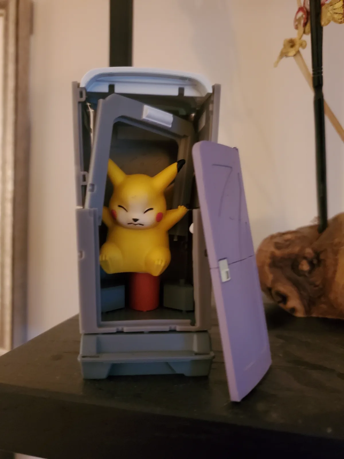 A yellow Pikachu toy with closed eyes and a smug expression is sitting inside an open, grey plastic Poké Ball toy, with the…