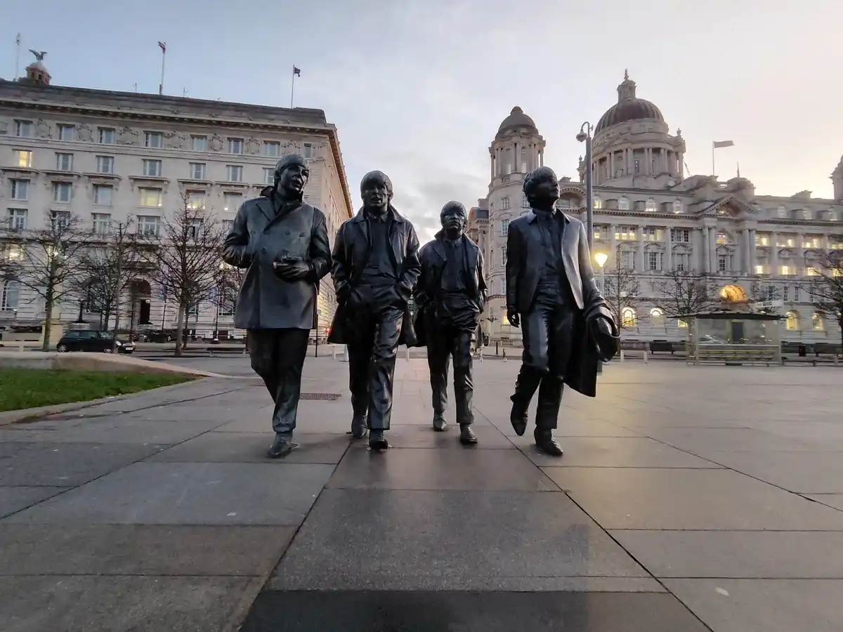 A photograph of four bronze statues of The Beatles walking in a line on a paved plaza, with grand classical buildings and…
