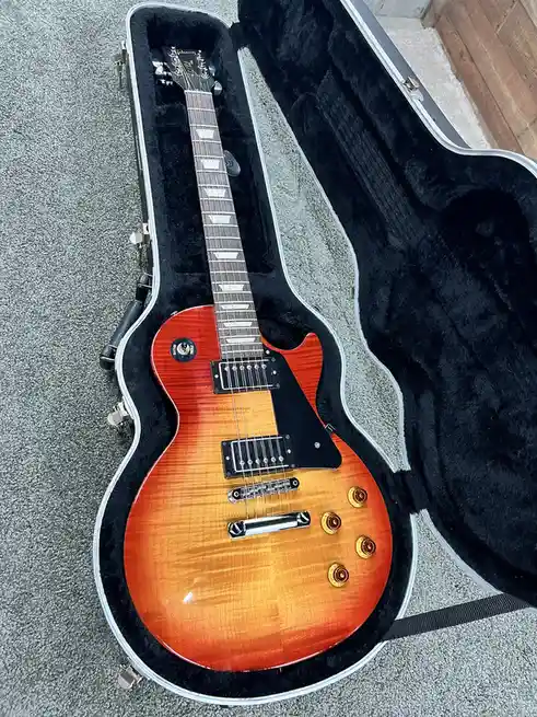 A Gibson Les Paul electric guitar with a sunburst finish sits inside its open black hardshell case, resting on a gray…