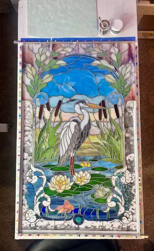 A stained glass artwork depicting a heron standing in water with lily pads and lotus flowers, surrounded by green reeds and…
