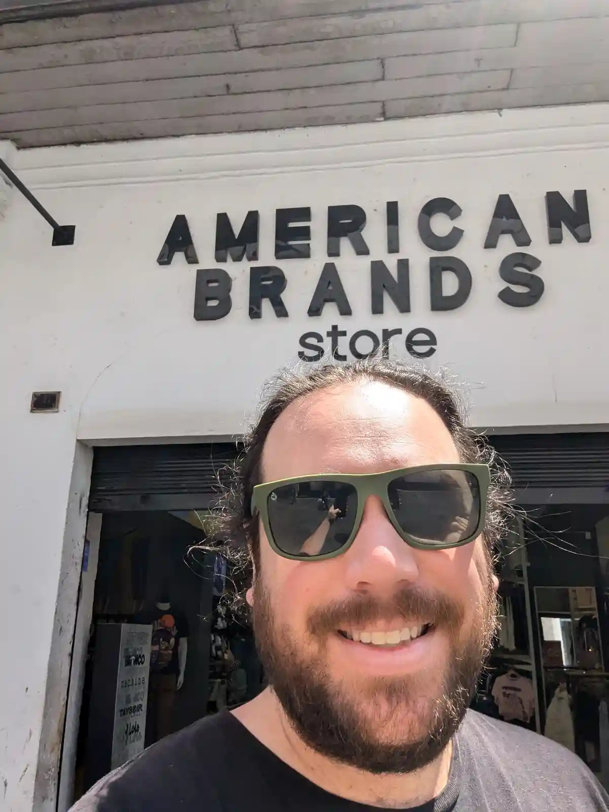 A man with a beard and green sunglasses smiles for a selfie in front of a store with a sign that reads 'AMERICAN BRANDS…