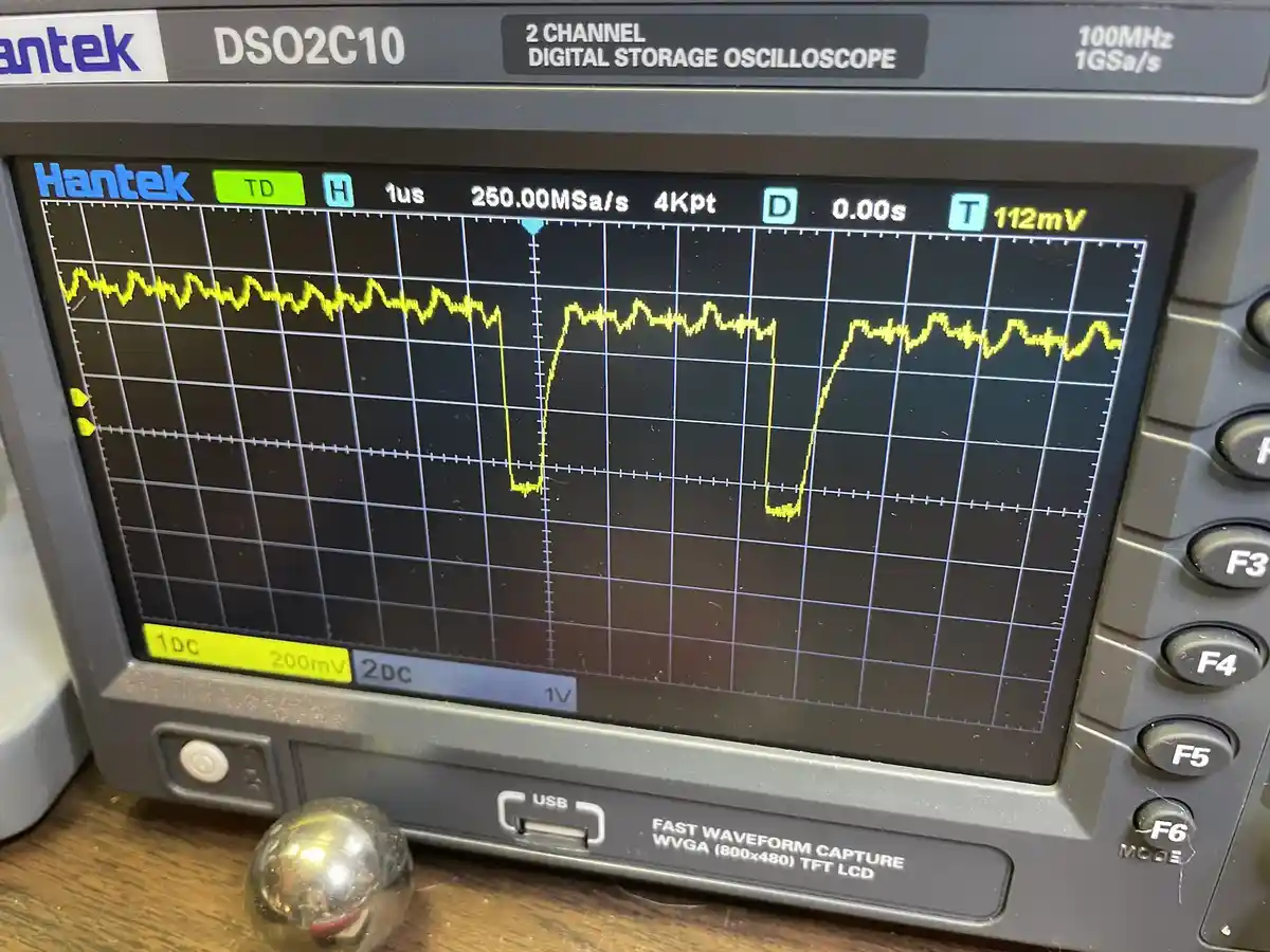 A close-up of a Hantek DSO2C10 digital storage oscilloscope displaying a jagged yellow waveform with two distinct…