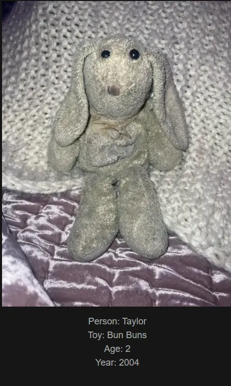 A gray, worn-out stuffed bunny toy with long floppy ears sits on a textured purple fabric, positioned against a…