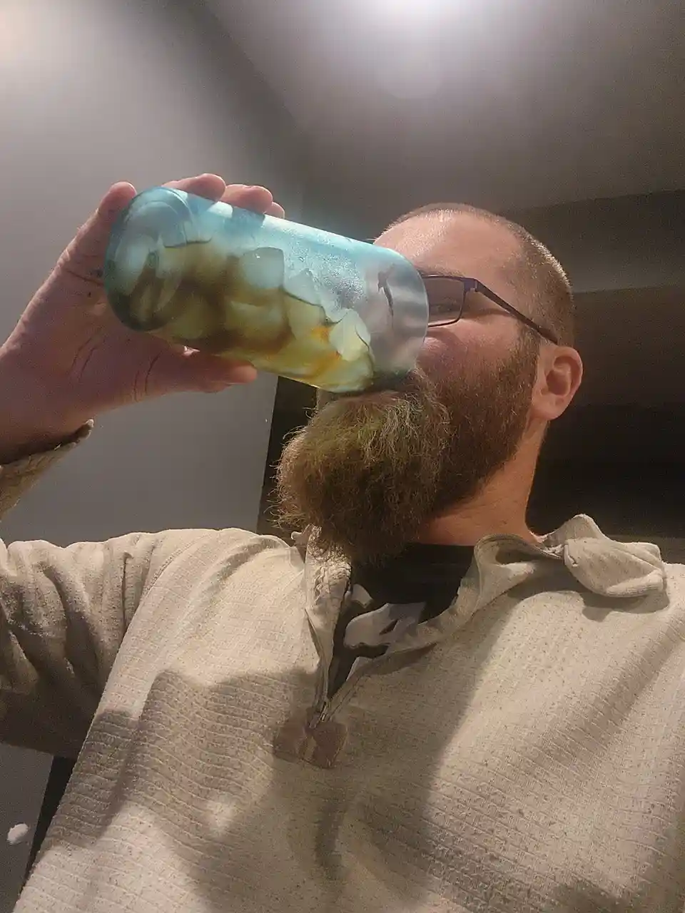 A man with glasses and a full beard is holding a large blue tumbler with ice and a dark liquid to his mouth as if drinking…