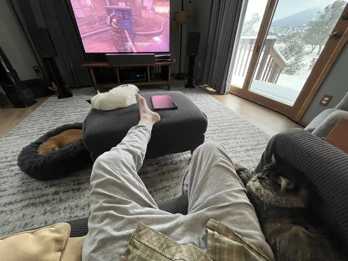 A person in pajamas lounges on a couch with their feet up on a grey ottoman, surrounded by three cats. One white cat sleeps…