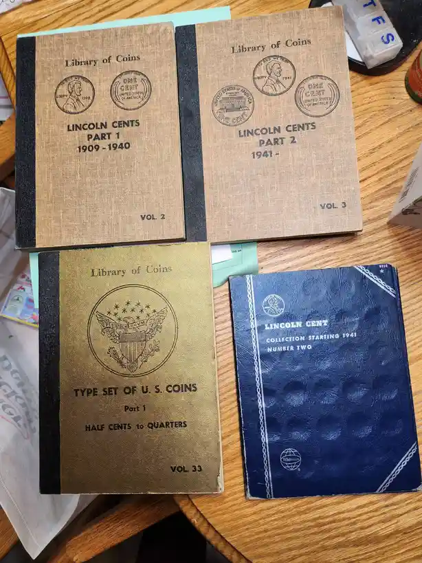 Four coin collecting albums are arranged on a wooden table. Two tan 'Library of Coins' binders cover Lincoln Cents, split…