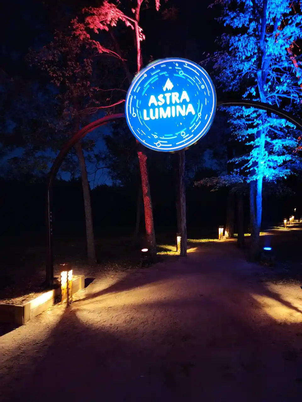 A nighttime photo of a glowing blue circular sign reading 'ASTRA LUMINA' suspended between two trees on a path, with trees…