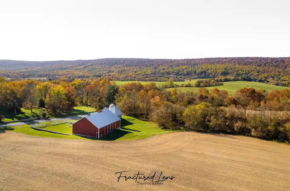 An aerial photo shows a red barn with a white silo nestled in a scenic autumn landscape with colorful trees and rolling…