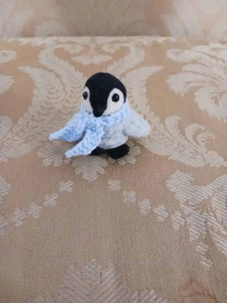 A small, handmade knitted penguin plushie with a black head, white face, and black feet sits on a beige patterned fabric…