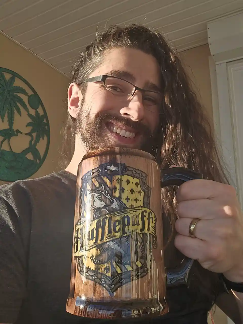 A smiling man with long hair and glasses holds up a large wooden mug featuring the Hufflepuff house crest from Harry Potter…