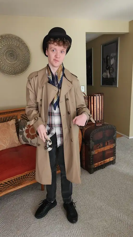 A young man with curly hair wears a black bowler hat, a beige trench coat, a plaid shirt, and a patterned scarf. He holds a…