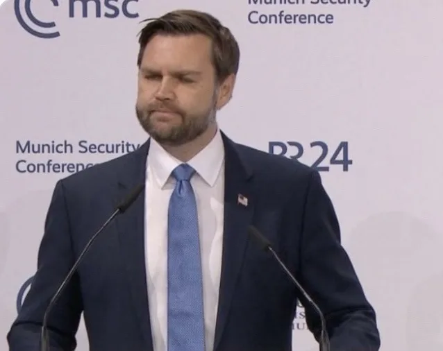 A man in a dark suit and blue tie stands at a podium with two microphones, looking down with a weary expression. Behind him…