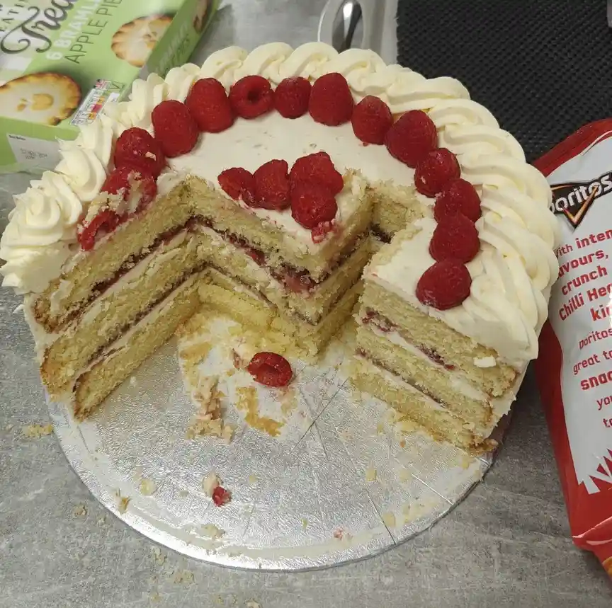 A partially eaten multi-layered cake with white frosting and raspberries on top sits on a silver cake board, with a slice…