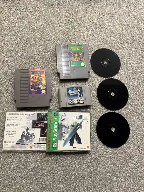 An overhead shot displays four video game cartridges and three black CDs arranged on a gray carpet. The cartridges are for…