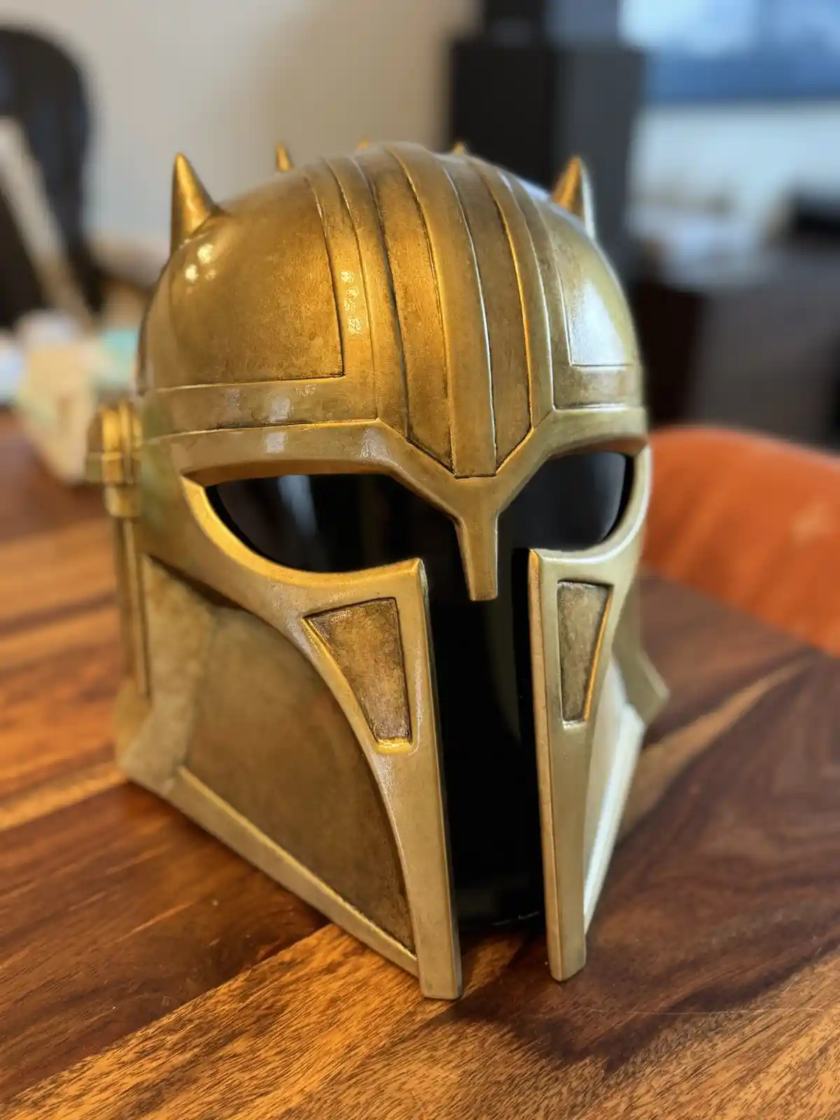 A golden Mandalorian helmet, resembling The Mandalorian's iconic armor from Star Wars, sits on a wooden table. The helmet…
