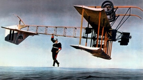 A man in a dark uniform with red trim hangs from the wing of a vintage biplane flying low over water. The aircraft has a…