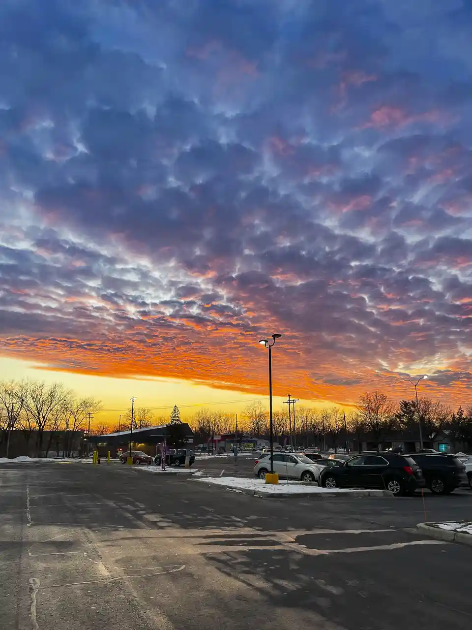 A photograph of a parking lot at dusk, with cars parked and snow patches on the ground, under a dramatic sky filled with…