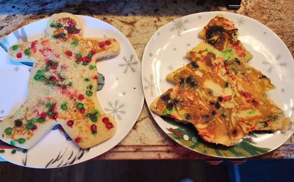 Two plates hold pancakes shaped like a gingerbread man and a Christmas tree, both decorated with red and green sprinkles…