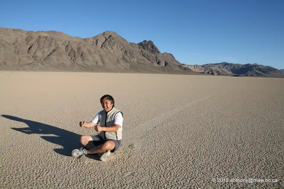 a man sits cross-legged on a vast, cracked desert plain with mountains in the background, smiling broadly with clenched…