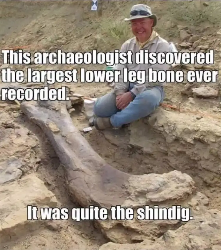 A smiling man in a hat and outdoor clothing sits beside a massive, unearthed fossilized bone in a dirt excavation site. The…