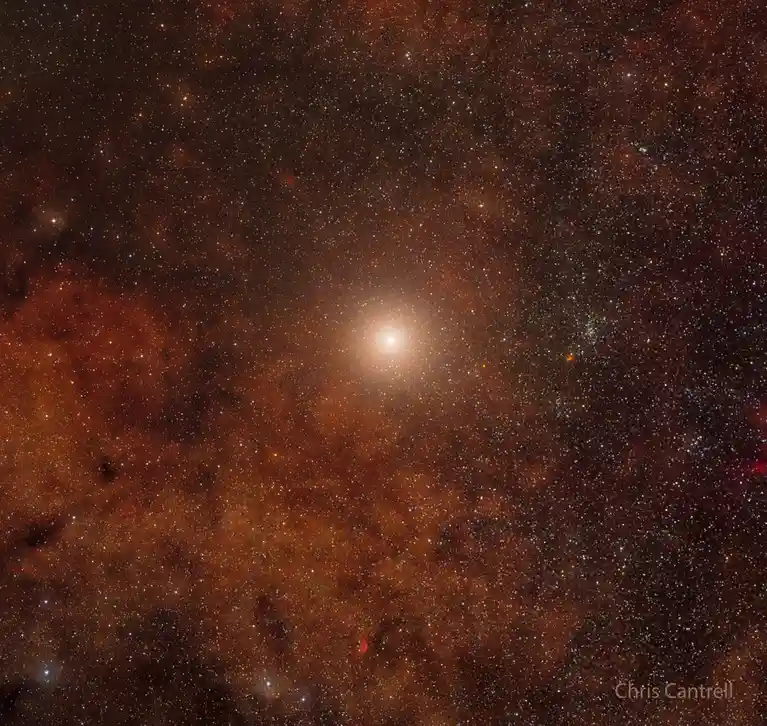 A deep space photograph shows a bright central star surrounded by a dense field of stars and reddish-brown cosmic dust…