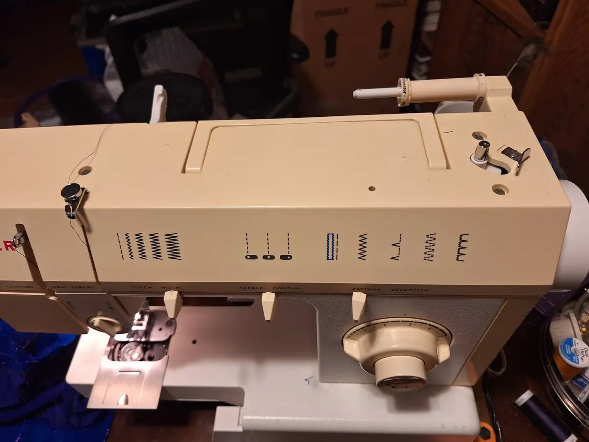 A close-up photo of a beige sewing machine with visible stitch pattern dials and controls, showing a thread spool on top and…