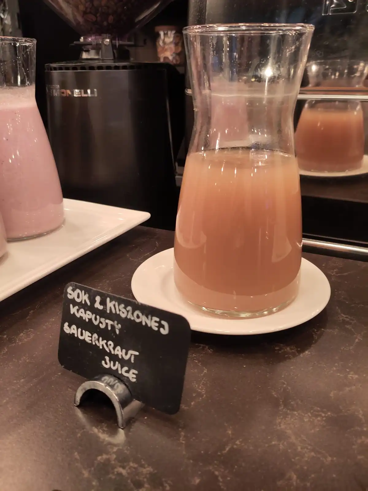A glass carafe filled with pinkish-brown liquid sits on a white saucer on a dark countertop, next to a small black…