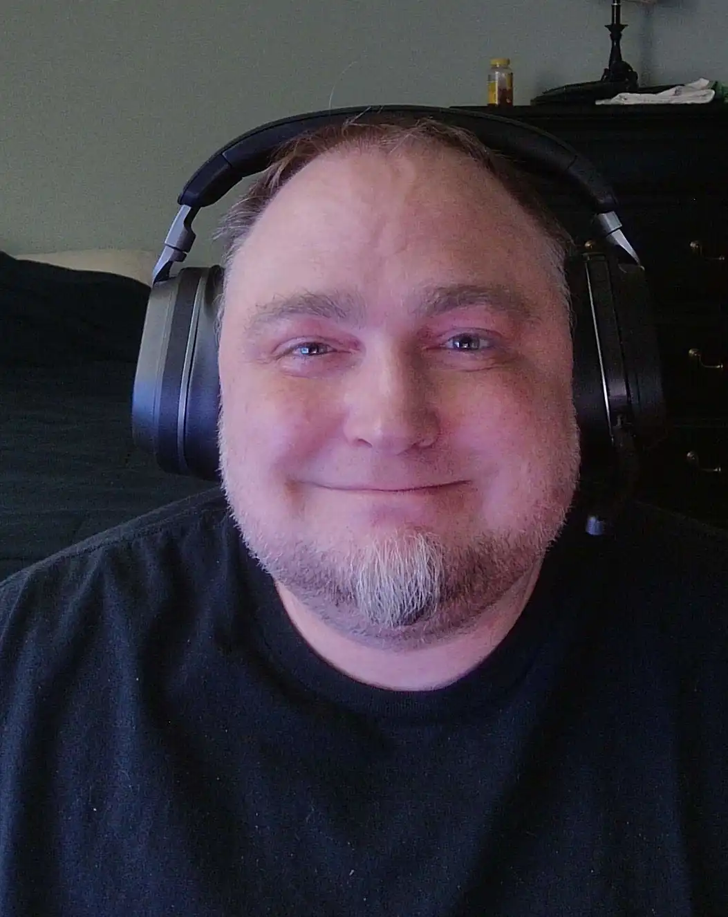 A man with a receding hairline and goatee, wearing large black over-ear headphones, smiles faintly while looking at the…