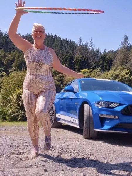 A woman in a shiny, patterned one-piece outfit stands outdoors next to a blue Ford Mustang, holding a colorful hula hoop…
