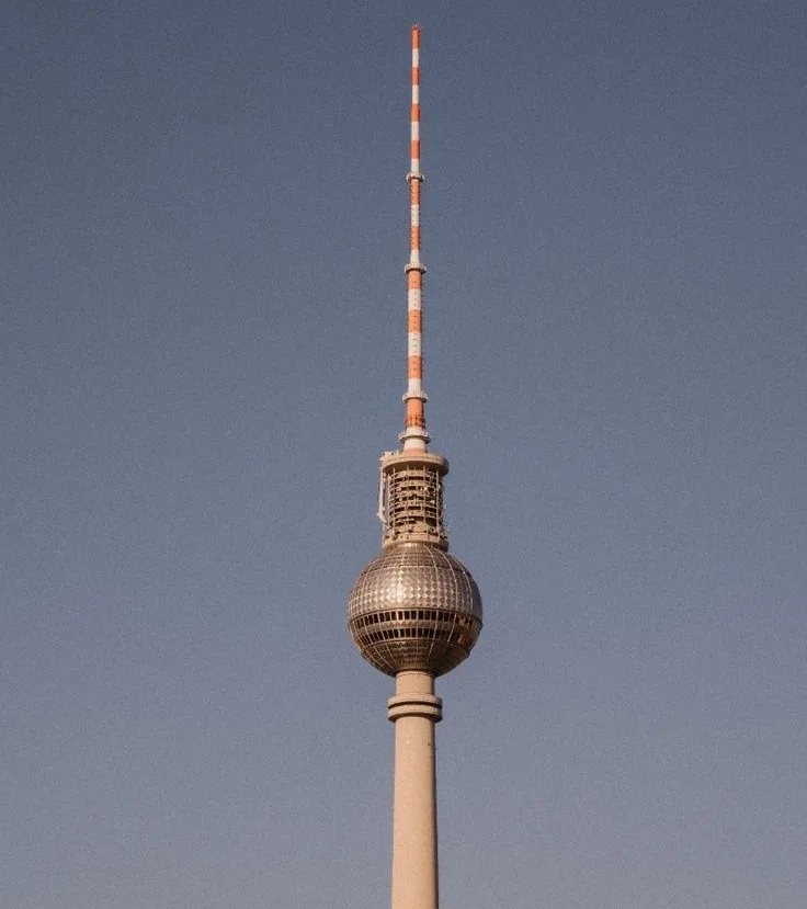 A low-angle photo shows the top section of the Berlin TV Tower against a clear blue sky, featuring its distinctive spherical…
