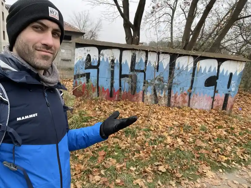 A man in a blue and black Mammut jacket and black beanie stands outdoors in autumn, gesturing toward a rusty metal shed…