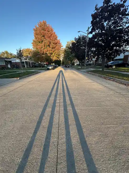 A low-angle photo of a quiet suburban street at sunrise or sunset, with long, distinct shadows of two people stretching down…