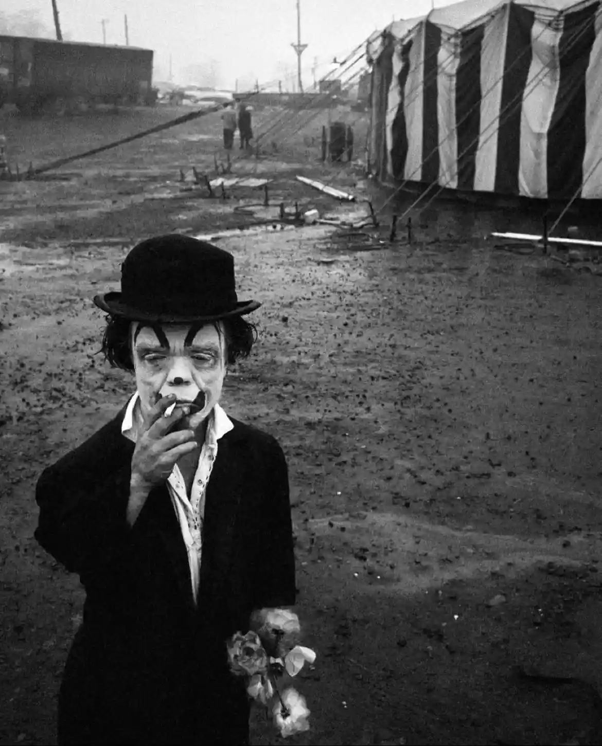 A black-and-white photograph shows a man with clown makeup, wearing a bowler hat and holding a bouquet of flowers, smoking a…