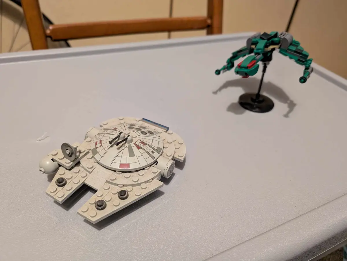 A white LEGO Millennium Falcon model sits on a white table, with a green LEGO starfighter on a black stand in the…