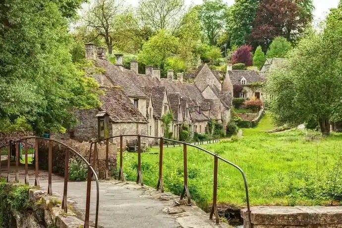A picturesque view of a quaint village with stone cottages and a curved metal bridge over a grassy area. The overlaid text…