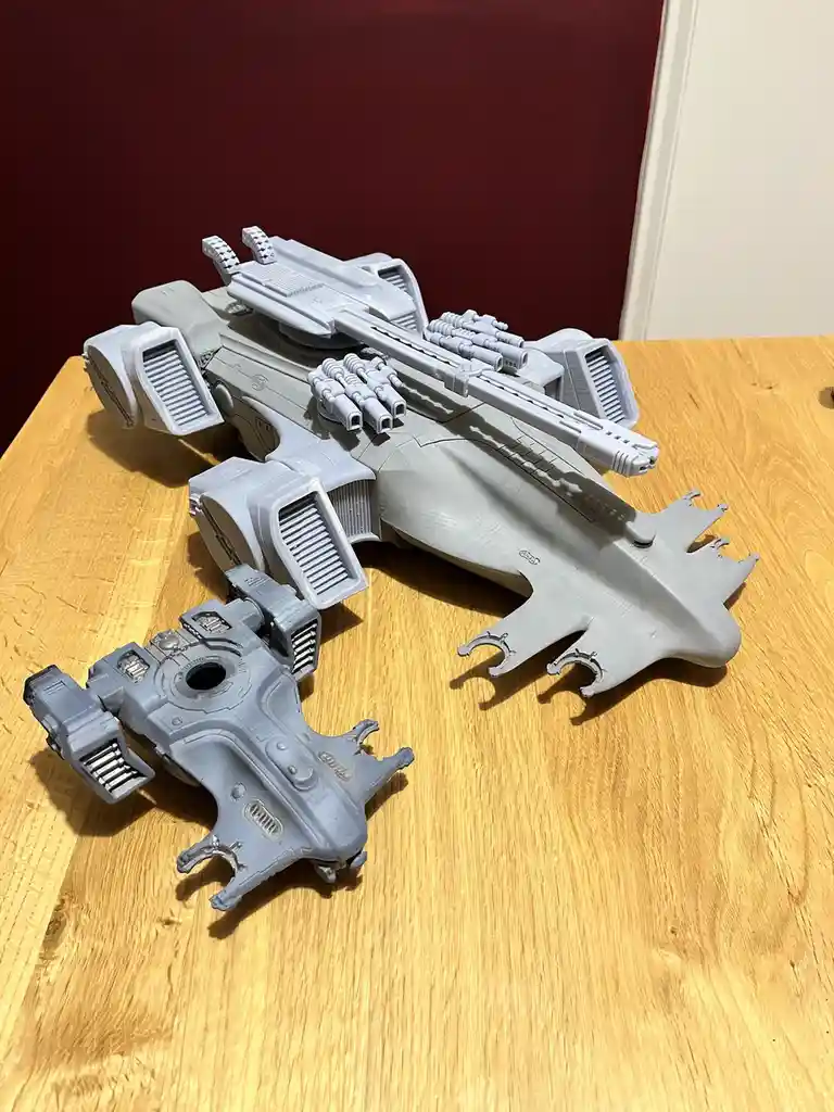 A large, gray 3D-printed spaceship model sits on a wooden table, with a smaller, detached section lying nearby. The model…