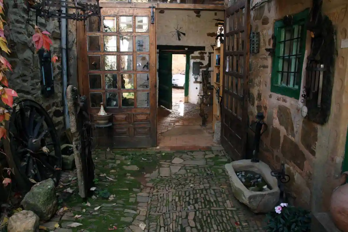A rustic courtyard with stone walls, a cobblestone path, and an open wooden door leading inside. Decorative items like a…