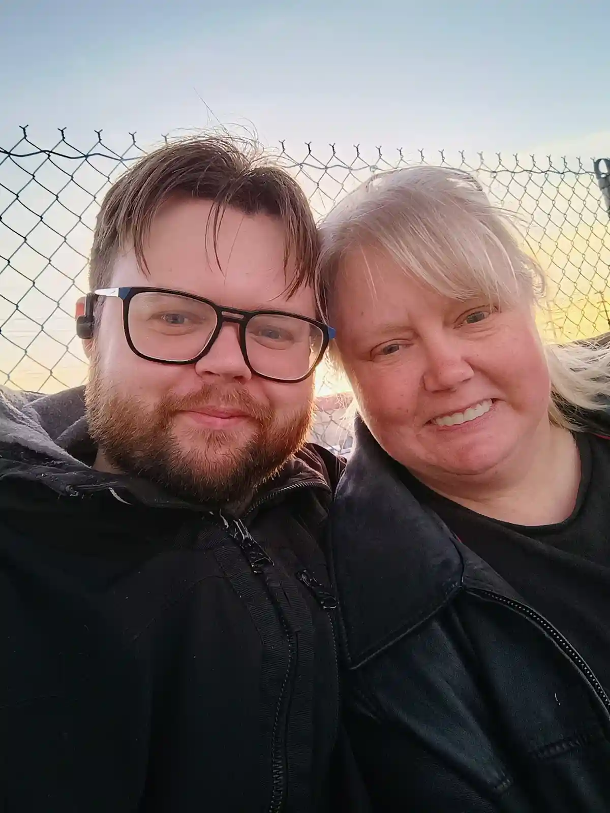 A selfie of two people, a man with glasses and a beard and a woman with blonde hair, standing close together in front of a…
