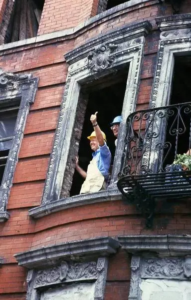 Two construction workers in hard hats and work clothes stand in an ornate, broken window of a red brick building, one giving…