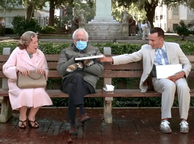 Three people sit on a park bench; a man in a beige suit offers a box of donuts to a man in a green jacket wearing a blue…