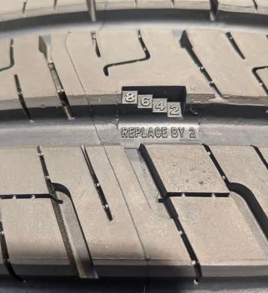 A close-up of a worn tire tread shows embossed numbers '8 6 4 2' above the phrase 'REPLACE BY 2', creating a visual pun…